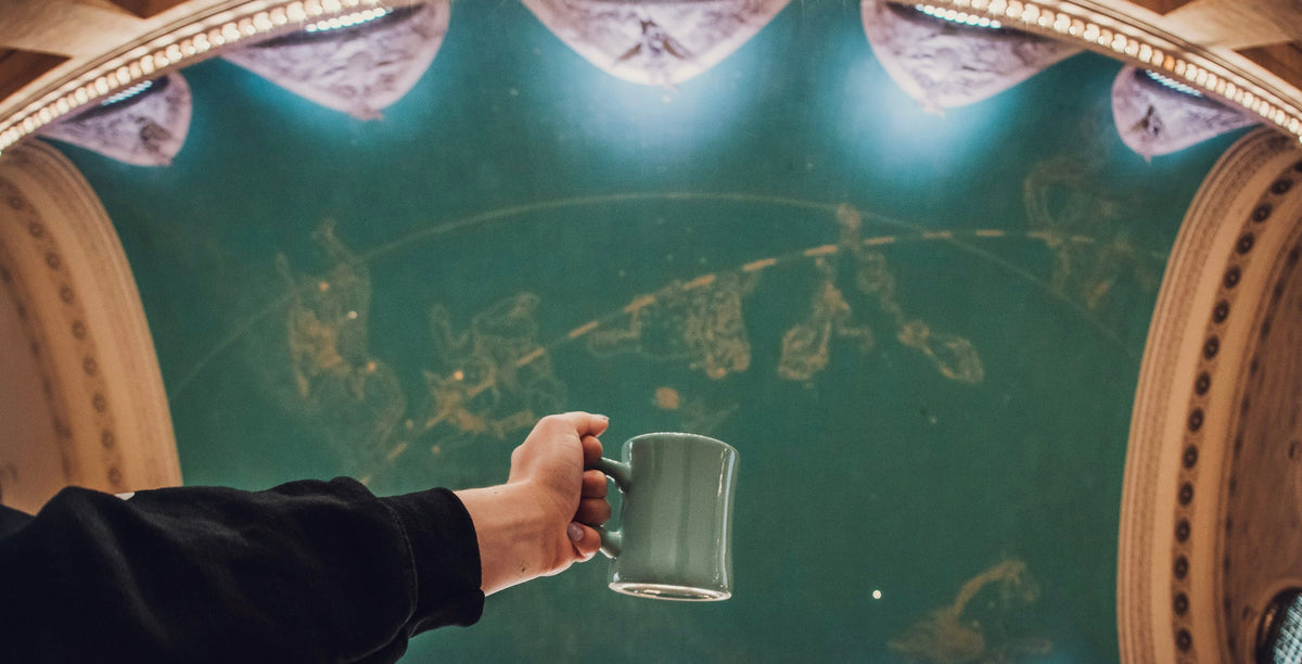 A mug of coffee held up under the ceiling of Grand Central Terminal